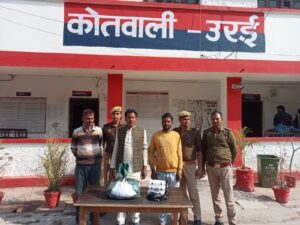 जालौन पुलिस की गिरफ्त में खड़े तीन गांजा तस्कर, मेज पर बरामद गांजा, नकदी, मोबाइल और पीछे खड़ी सफेद स्कॉर्पियो कार