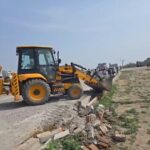 उरई के झांसी रोड पर अवैध प्लॉटिंग को ध्वस्त करता विकास प्राधिकरण का बुलडोजर और मौके पर उपस्थित पुलिस बल एवं अधिकारी