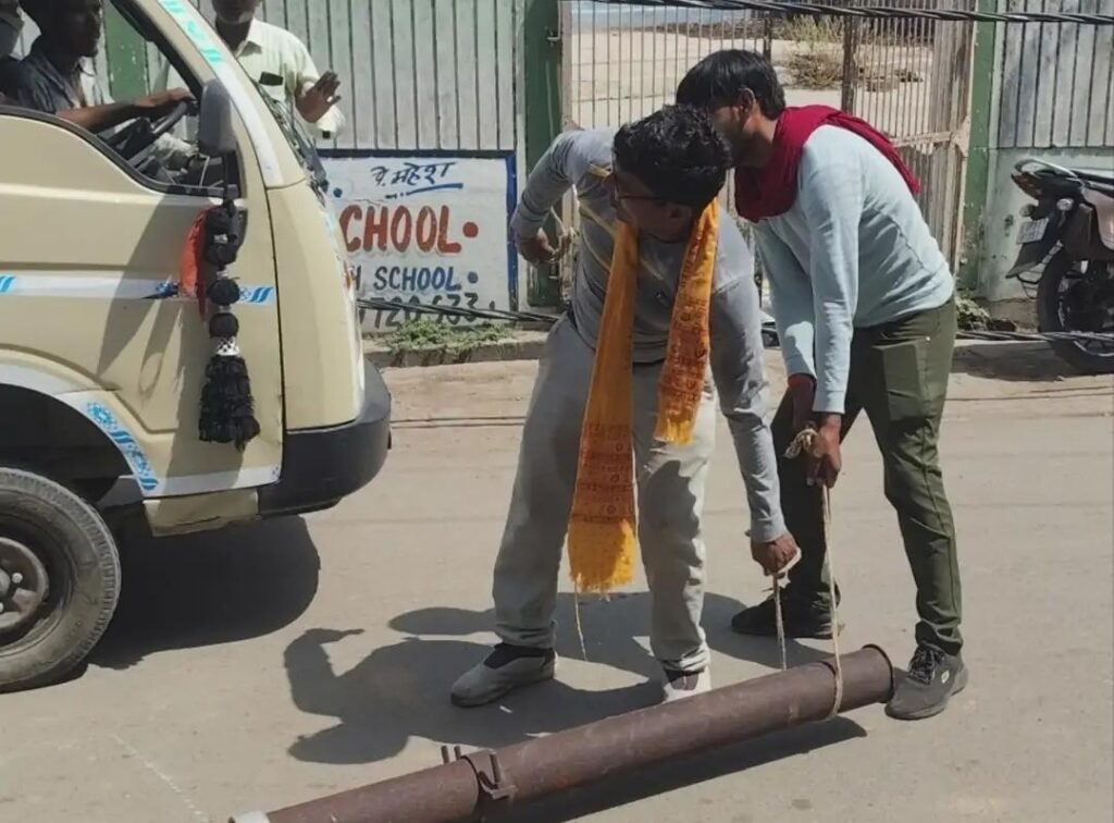 जालौन के कोंच में मुख्य मार्ग पर कार के ऊपर गिरा टूटा हुआ जर्जर बिजली का पोल, क्षतिग्रस्त कार और आस-पास जमा भीड़ का दृश्य