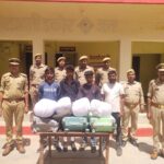 जालौन पुलिस की गिरफ्त में खड़े चार गांजा तस्कर, मेज पर बरामद 24 किलो अवैध गांजा, तीन ट्रॉली बैग और मोबाइल फोन रखे हुए।