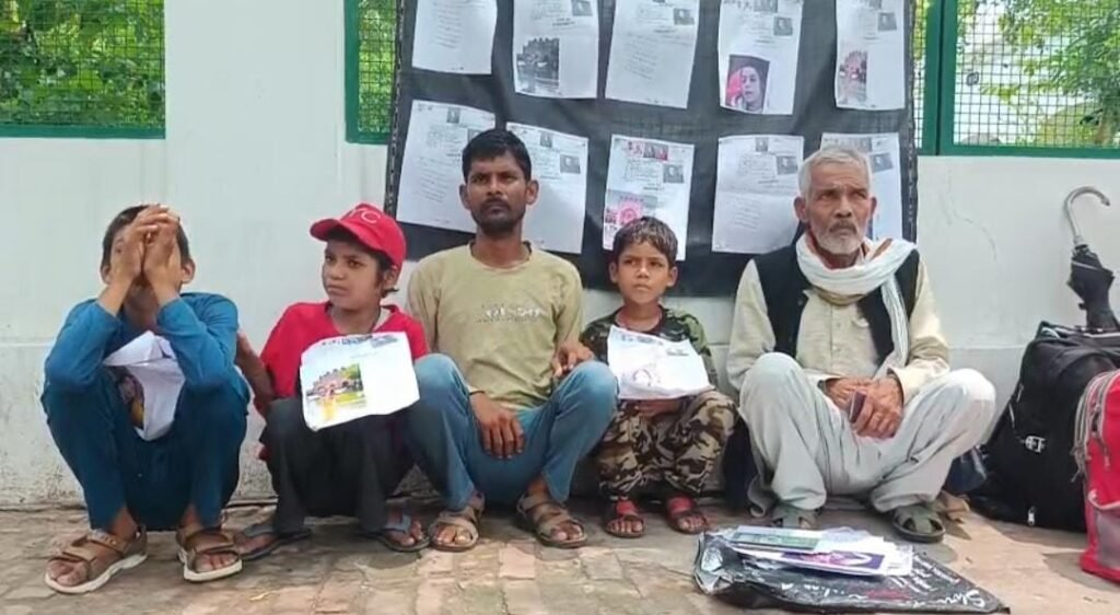 जालौन के जिलाधिकारी कार्यालय के बाहर अपनी तीन मासूम बच्चों के साथ जमीन पर धरने पर बैठा एक मजदूर पति, जो हाथ में पत्नी की तलाश की गुहार से जुड़ा पोस्टर लिए न्याय की मांग कर रहा है।